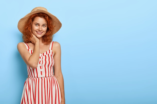 Free photo shot of happy redhaired woman dressed in fashionable summer outfit, straw hat, has charming smile, natural beauty, stands against blue  wall with blank space for your promotion.