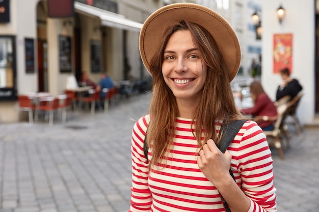 Shot of lovely female smiles gladfully, wears hat and striped jumper, being in good mood as strolls across city