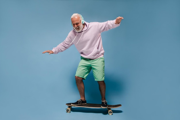 Shot of man in green shorts and hoodie riding skate board Positive guy with beard and headphones posing on isolated background