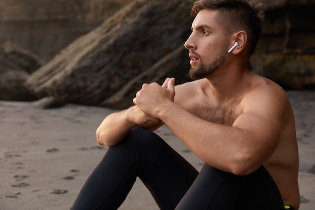 Free Photo shot of pensive caucasian male athlet keeps hands together, sits at sandy beach, wears leggings