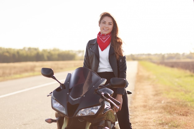 Shot of pleasant looking woman biker sits on fast black motorbike, wears red stylish bandana and leather jacket, travels alone