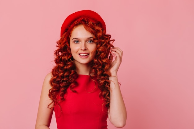 Shot of positive lady with blue eyes and red curls on pink space. Girl in bright headdress and red top looking at camera.