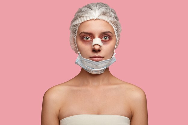 Shot of serious woman wears medical hat and mask, has rhinoplasty, preapres for blepharoplasty, has bruises around eyes, looks seriously at camera,