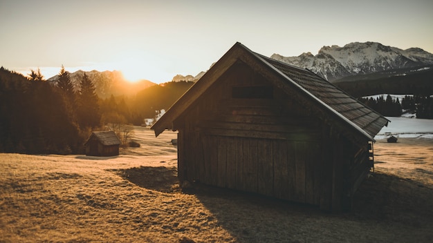 Free photo shot of a small wooden house with dry grass around it during sunset with mountains in the backgro