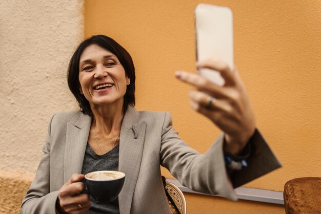 Shot of smiling fairskinned adult woman using her mobile phone while having cup of coffee outside Gadget technology concept