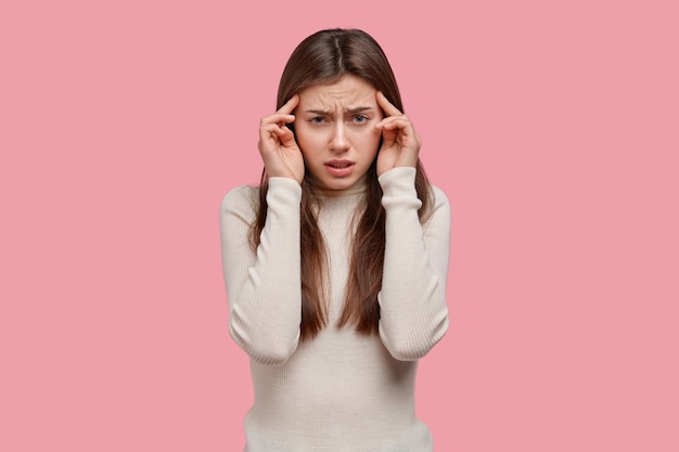 Free photo shot of sorrowful young woman keeps fingers on temples, has headache, feels exhausted and displeased, tries to think on solving problem