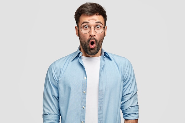 shot of terrified unshaven man stares with eyes full of disbelief, being surprised by curious revelation, wears blue shirt, isolated over white wall. People, reaction, facial expressions