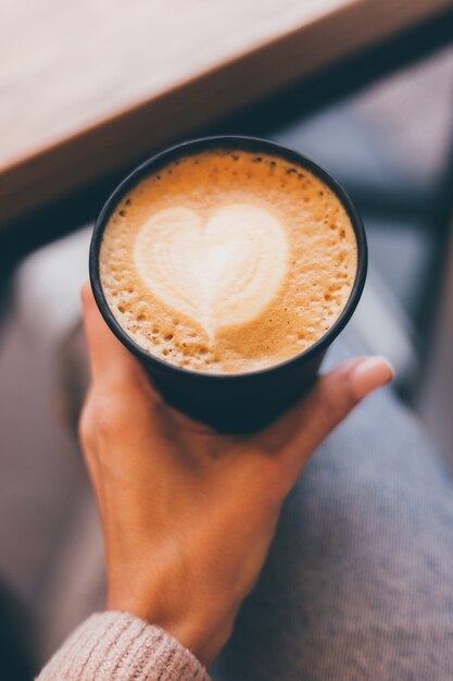 Shot of woman hands hold cup of hot coffee with heart design made of foam.