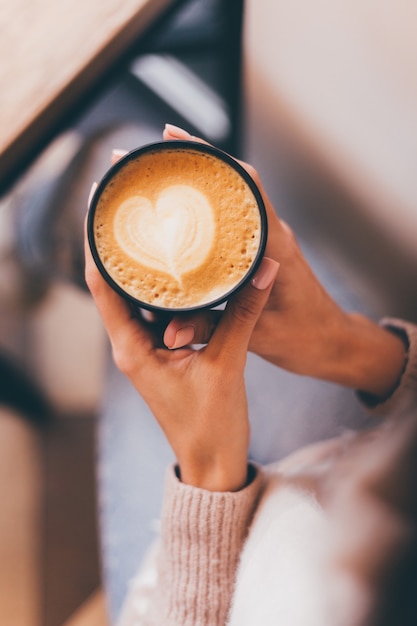 Shot of woman hands hold cup of hot coffee with heart design made of foam.