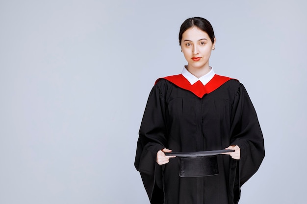 Shot of young student in gown holding cap against wall. High quality photo