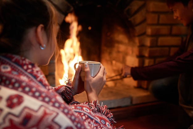 Free Photo over the shoulder shot of woman warming her hands on the mug of hot tea sitting at the fireplace, her boyfriend dealing with charcoal