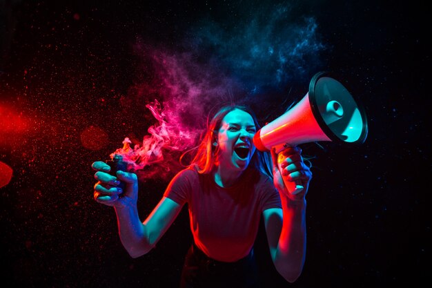 Shouting with megaphone. Young woman with smoke and neon light on black background. Highly tensioned, wide angle, fish eye view. 