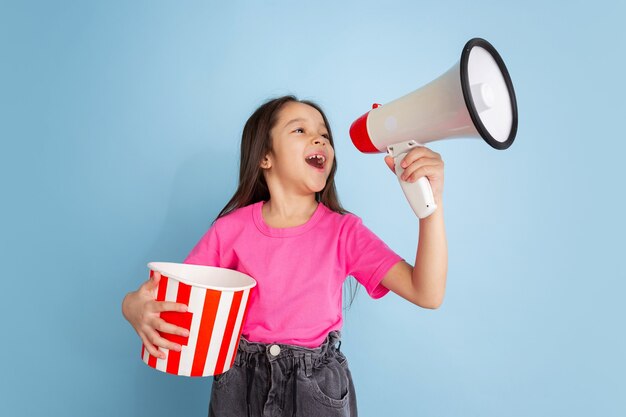 Shouting with popcorn. Caucasian little girl's portrait on blue  wall. Beautiful female model in pink shirt. Concept of human emotions, facial expression, youth, childhood.