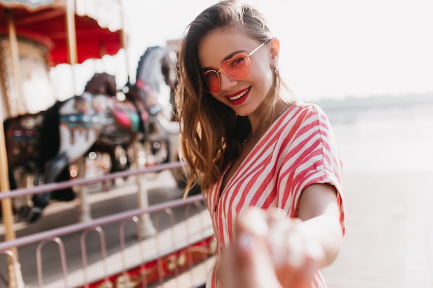 Shy beautiful girl in heart sunglasses standing near carousel. Outdoor portrait of blithesome woman in striped attire smiling