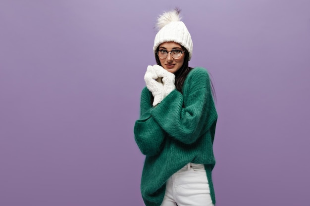 Free Photo shy cute woman in stylish woolen green sweater and eyeglasses poses on purple background pretty brunette girl in hat and gloves looks into camera