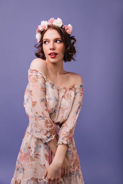 Shy european lady in romantic dress looking around. Well-dressed curly girl with flowers on her head posing.