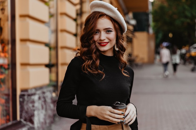 Free photo shy french woman with long hair posing outdoor. adorable red-haired lady standing on the street with cup of coffee.