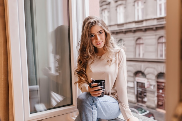 Shy woman in jeans sitting on window sill and posing with interest