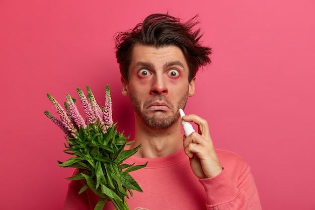 Sick allergic young man drips nose with nasal drops, has red eyes and nose, allergy on plant, symptoms of rhinitis or hay fever, stares , poses against pink wall, reacts on pollen