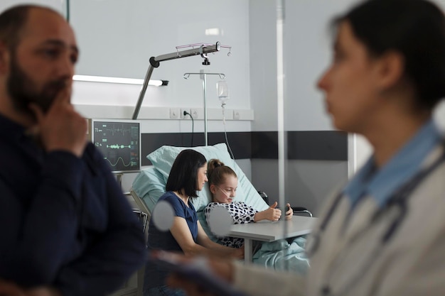 Sick girl playing games on smartphone with mother while father talking with expert pediatrist. Hospitalized ill little kid watching cartoons on mobile phone while resting in hospital pediatric ward