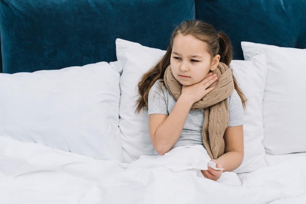 Sick girl sitting on bed with scarf around her neck suffering from sore neck