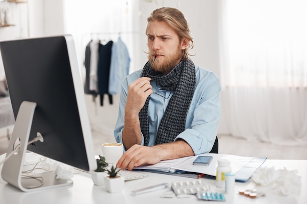 Free photo sick ill bearded man sits in front of computer, tries to concentrate on work, holds spectacles in hand. exahausted office worker tired, has sedentary lifestyle, isolated against office background.