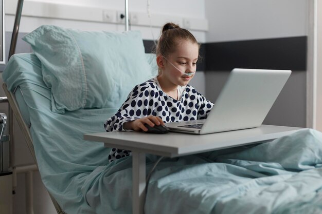 Sick little girl playing games on laptop while hospitalized in healthcare clinic pediatric ward room. Ill kid resting on patient bed while enjoying gaming on modern computer.