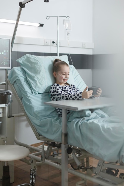 Free photo sick little girl playing on smartphone while resting on patient bed in pediatric clinic treatment room. joyful young patient enjoying mobile phone entertainment while sitting in clinic recovery ward.