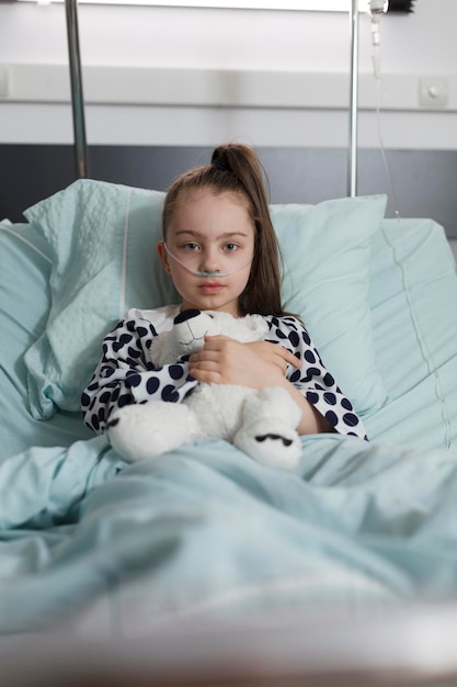 Free photo sick little girl under treatment wearing oxygen tube while holding plush bear toy. ill kid resting in children healthcare facility patient bed alone while having teddy bear.