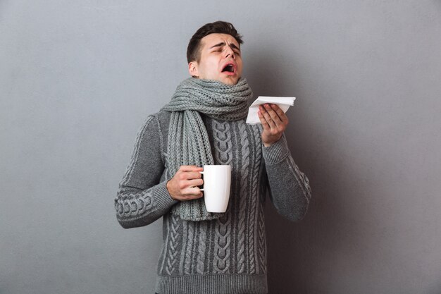 Sick Man in sweater and scarf sneezes while holding cup of tea