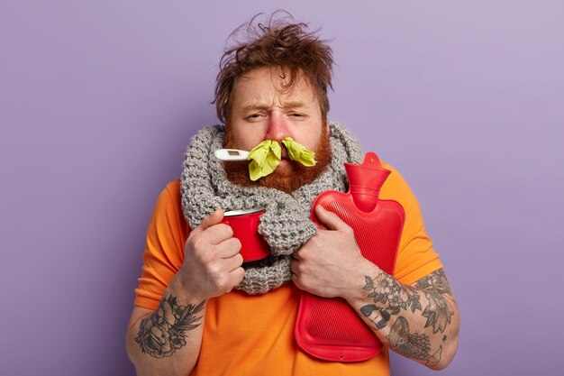 Sick man in warm clothes with thermometer, cup and water bag