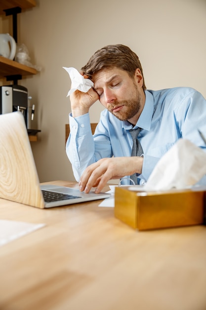 Sick man with handkerchief sneezing blowing nose while working in office