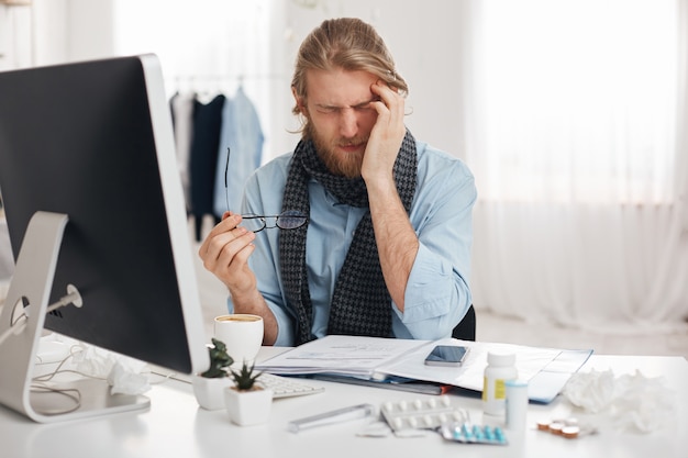 Sick and tired bearded male student or office worker has sleepy expression, runs temple because of feeling sick, surrounded by pills and drugs, tries to concentrate and finish work quicker