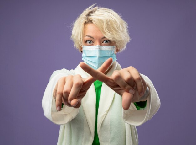 Sick unhealthy woman with short hair in facial protective mask making stop sign crossing index fingers standing over purple wall