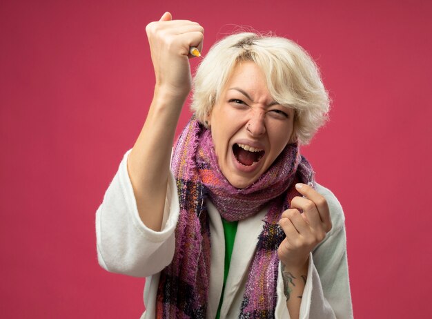 Sick unhealthy woman with short hair in scarf feeling unwell holding ampoule showing syringe shouting with aggressive expression crazy mad standing over pink background