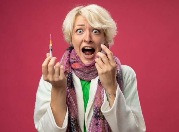 Sick unhealthy woman with short hair in scarf feeling unwell holding ampoule and syringe shouting in panic standing over pink wall