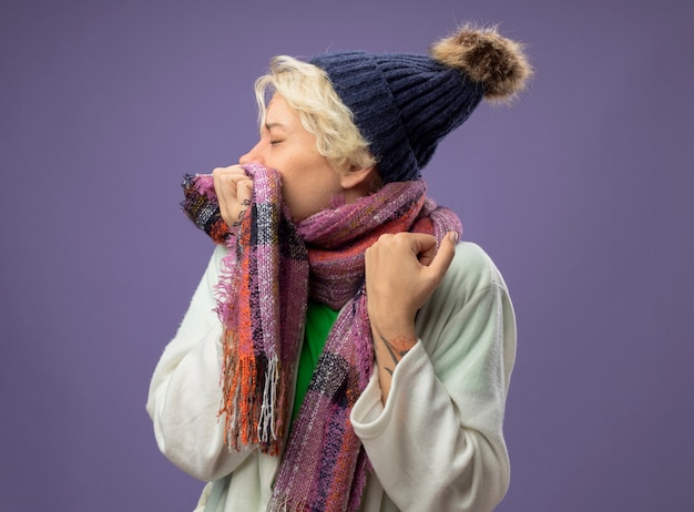 Free photo sick unhealthy woman with short hair in warm hat and scarf feeling unwell coughingover purple wall