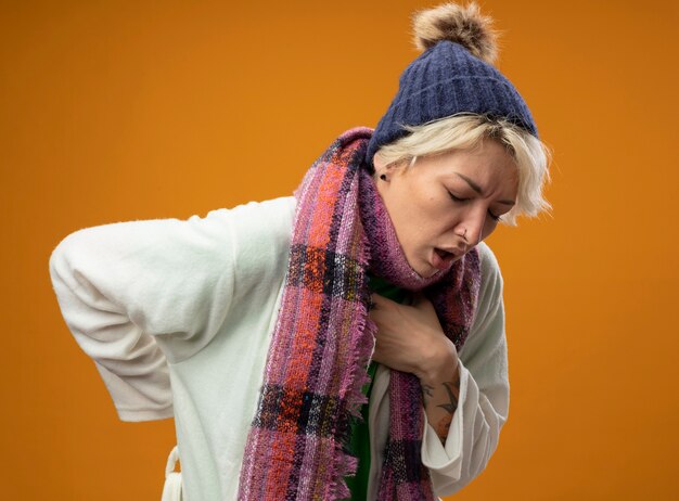 Sick unhealthy woman with short hair in warm scarf andhat feeling unwell coughing suffering from cold standing over orange background