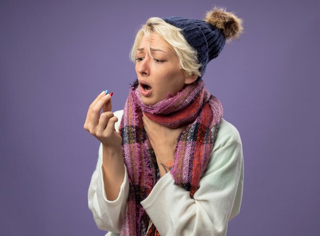 Sick unhealthy woman with short hair in warm scarf and hat feeling unwell holding pills looking at them worried standing over purple background