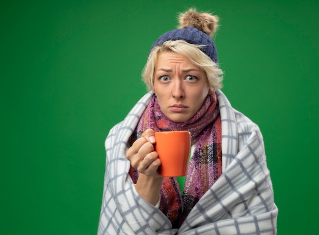 Sick unhealthy woman with short hair in warm scarf and hat feeling unwell wrapped in blanket holding cup of tea  standing over green wall