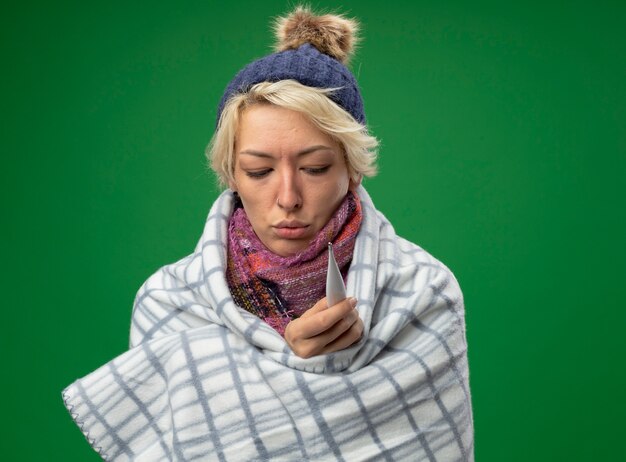 Sick unhealthy woman with short hair in warm scarf and hat feeling unwell wrapped in blanket holding thermometer standing over green background