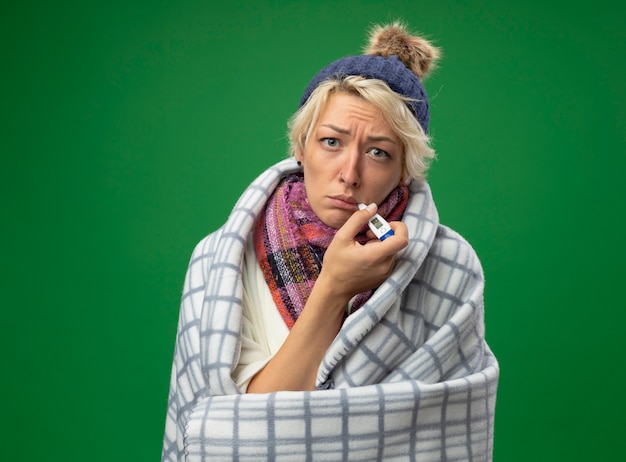 Free photo sick unhealthy woman with short hair in warm scarf and hat feeling unwell wrapped in blanket with thermometer in her mouth standing over green background