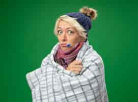 Free photo sick unhealthy woman with short hair in warm scarf and hat feeling unwell wrapped in blanket with thermometer in her mouth standing over green background