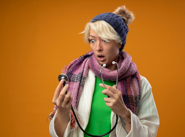 Free photo sick unhealthy woman with short hair in warm scarf and hat with stethoscope looking at it confused and amazed standing over orange background