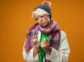 Free photo sick unhealthy woman with short hair in warm scarf and hat with stethoscope looking at it with sad expression standing over orange wall