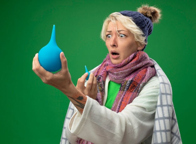 Free photo sick unhealthy woman with short hair in warm scarf and hat wrapped in blanket holding enema looking at it beingsurprised standing over green background