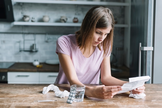 Free photo sick woman looking at prescription with blister pack and glass of water on wooden desk