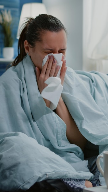 Sick woman using tissue to blow runny nose having cold. Unhappy adult feeling ill and drinking tea from cup and sitting on couch with blanket. Person with disease treatment and medicine