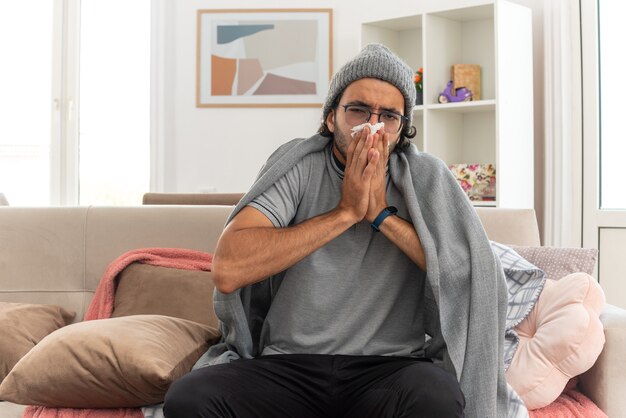 Sick young caucasian man in optical glasses wrapped in plaid wearing winter hat wipes his nose with tissue looking at camera sitting on couch at living room