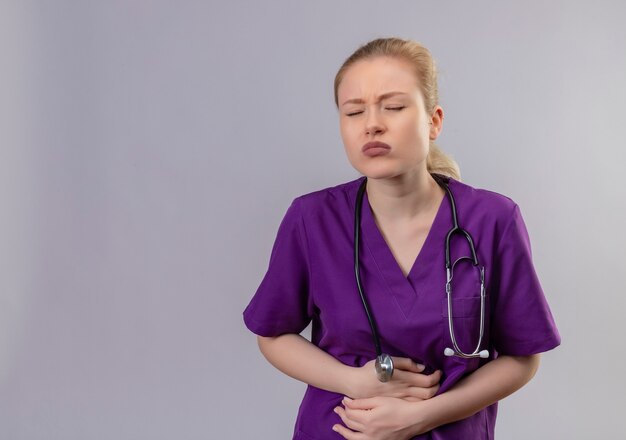 Sick young doctor wearing purple medical gown and stethoscope grabbed aching stomach on isolated white wall